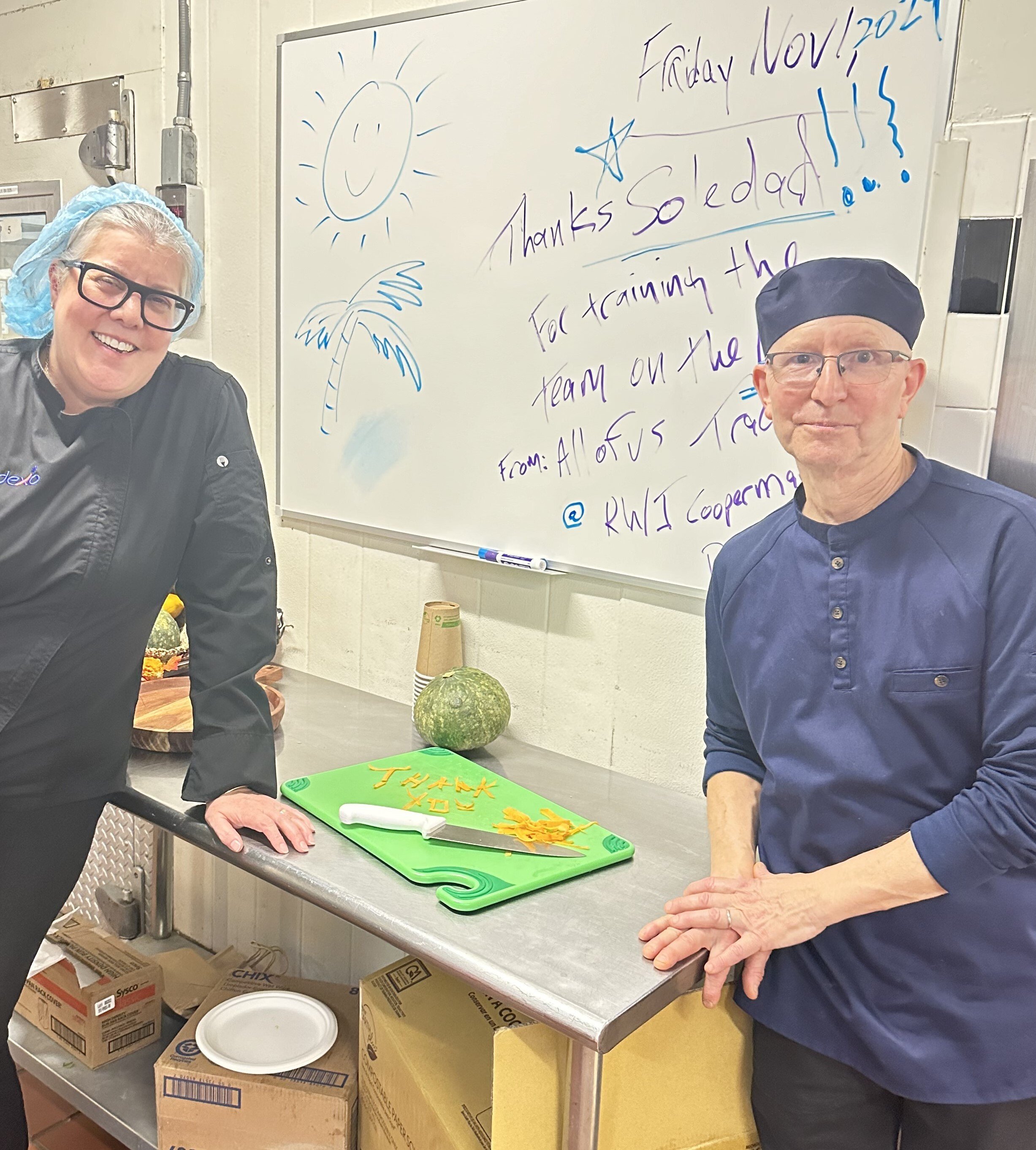 Kitchen whiteboard reading "Thanks Soledad for training the team" next to a cutting board with "thank you" spelled out in carrot peel